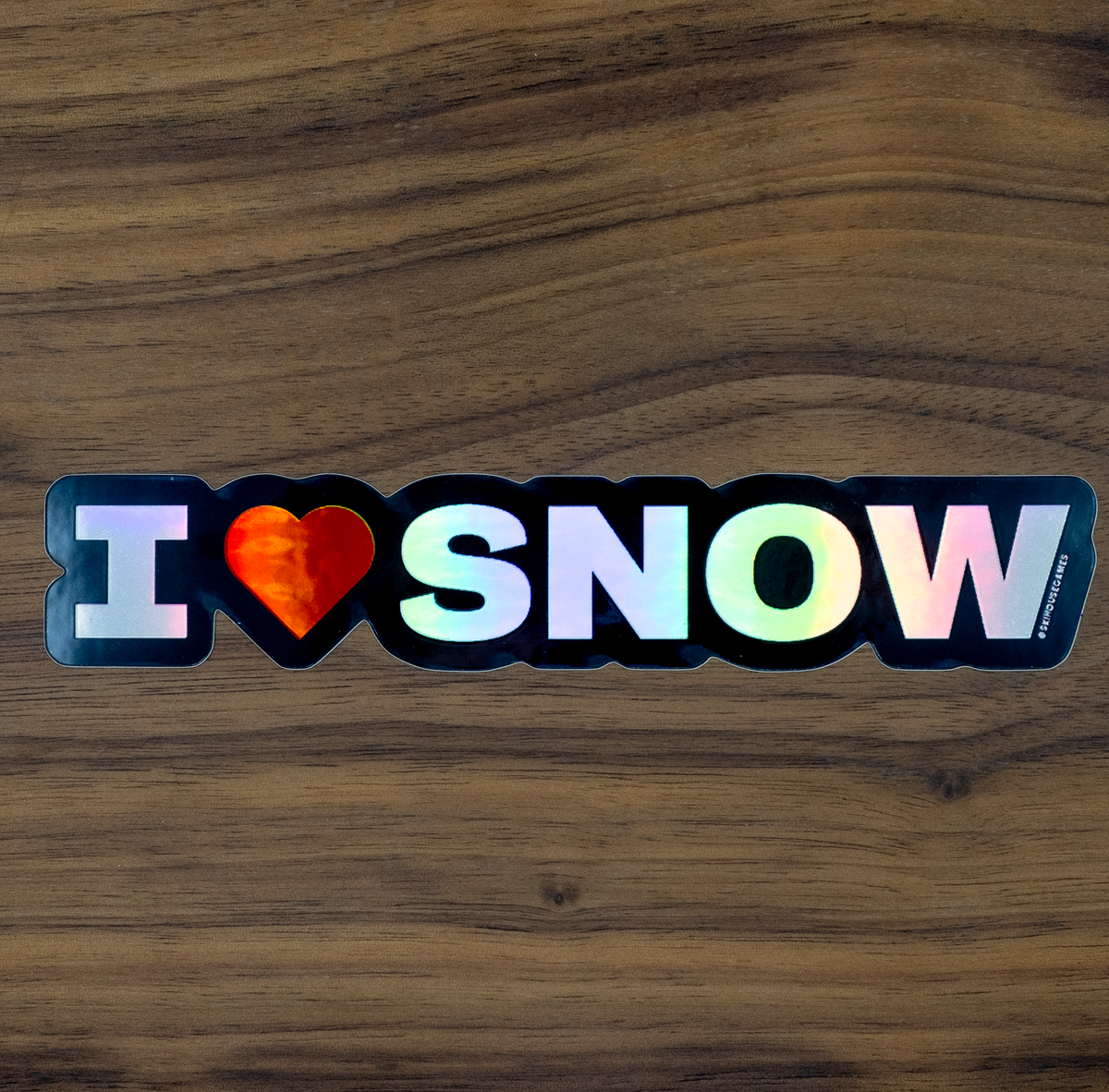 A die-cut sticker with the text 'I [heart] SNOW' in white and iridescent letters, placed on a dark wood background. The heart is filled with a red-orange flame pattern, and a small vertical brand mark 'SKIHOUSEGAMES' is visible on the bottom right edge of the sticker.